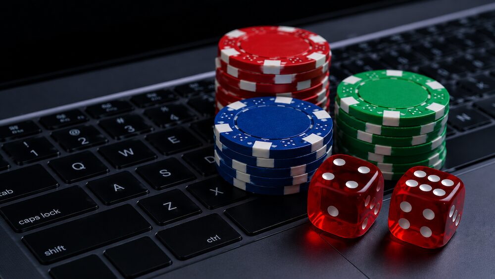Fotografía en primer plano de tres pilas de fichas de casino en colores rojo, azul y verde, dispuestas junto a un par de dados rojos translúcidos que descansan sobre el teclado negro de una computadora portátil.