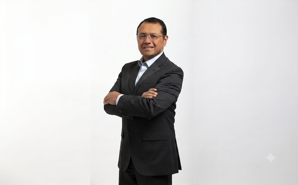 AMD potencia la IA: Retrato de un ejecutivo sonriente con anteojos y traje oscuro, de pie con los brazos cruzados frente a un fondo blanco liso y bien iluminado.
