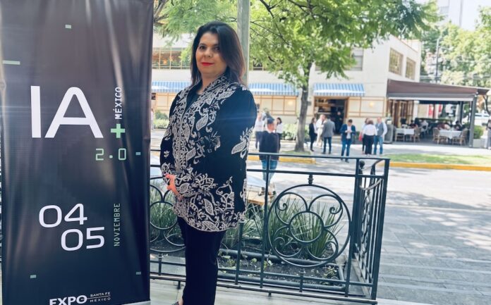 El liderazgo detrás de la estrategia nacional de inteligencia artificial Fotografía de una mujer de cabello oscuro vistiendo una chaqueta negra con bordados blancos, de pie en un espacio al aire libre junto a un pendón publicitario oscuro que anuncia el evento