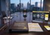 Norton alerta: 83% de los padres en México teme el uso de IA para crear contenido falso sobre sus hijos Fotografía de una elegante sala de juntas con vista panorámica a la ciudad al atardecer. Sobre la mesa de madera se encuentra un reporte impreso con el logotipo de Norton que destaca en su portada: "El 83% de padres teme que la IA cree imágenes o conversaciones falsas de sus hijos". A la derecha, hay un teléfono inteligente y un panel de acrílico transparente que muestra una infografía sobre protección infantil, reconocimiento facial y detección de seguridad.