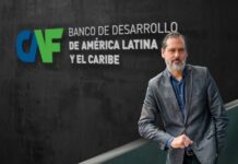 CAF revela cómo la transformación digital en América Latina redefine el futuro del canal TI Fotografía de Pablo Bello, con barba cana y vistiendo un saco gris sobre una camisa azul, posando de pie frente a un muro oscuro que exhibe el logotipo institucional de CAF, Banco de Desarrollo de América Latina y el Caribe.