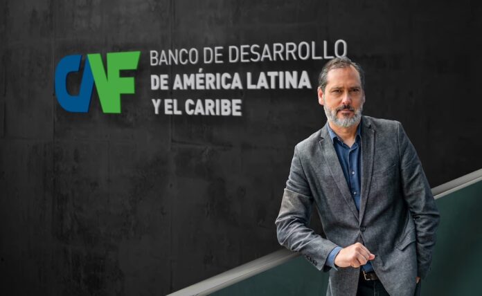 Fotografía de Pablo Bello, con barba cana y vistiendo un saco gris sobre una camisa azul, posando de pie frente a un muro oscuro que exhibe el logotipo institucional de CAF, Banco de Desarrollo de América Latina y el Caribe.