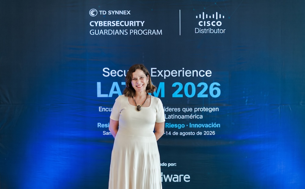 Retrato de Claudia Cincotto, sonriendo y vestida con un atuendo claro de falda plisada, de pie frente a un panel corporativo azul del programa Cybersecurity Guardians de TD SYNNEX.