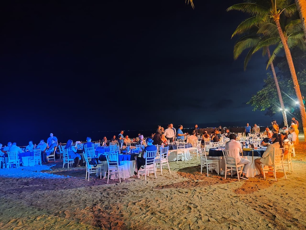 Fotografía nocturna al aire libre en la playa, mostrando a decenas de personas cenando y conversando alrededor de mesas iluminadas bajo palmeras, con el mar oscuro de fondo.