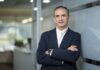 Hitachi Vantara fortalece al canal con soluciones de ciberresiliencia Fotografía de César Zurita, director comercial de Hitachi Vantara, posando en un entorno corporativo con los brazos cruzados. Viste un saco azul marino y camisa blanca sin corbata, apoyándose de forma relajada contra una pared de cristal en los pasillos de una oficina.