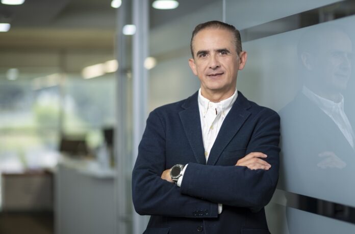 Fotografía de César Zurita, director comercial de Hitachi Vantara, posando en un entorno corporativo con los brazos cruzados. Viste un saco azul marino y camisa blanca sin corbata, apoyándose de forma relajada contra una pared de cristal en los pasillos de una oficina.