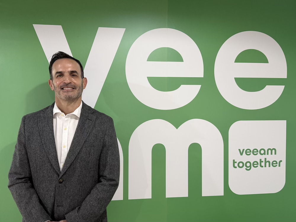 Fernando Zambrana asume el liderazgo estratégico de Veeam en México Fotografía de Fernando Zambrana sonriendo de frente a la cámara, vistiendo un saco gris texturizado y una camisa blanca sin corbata. Se encuentra de pie frente a un gran muro de color verde que muestra la palabra