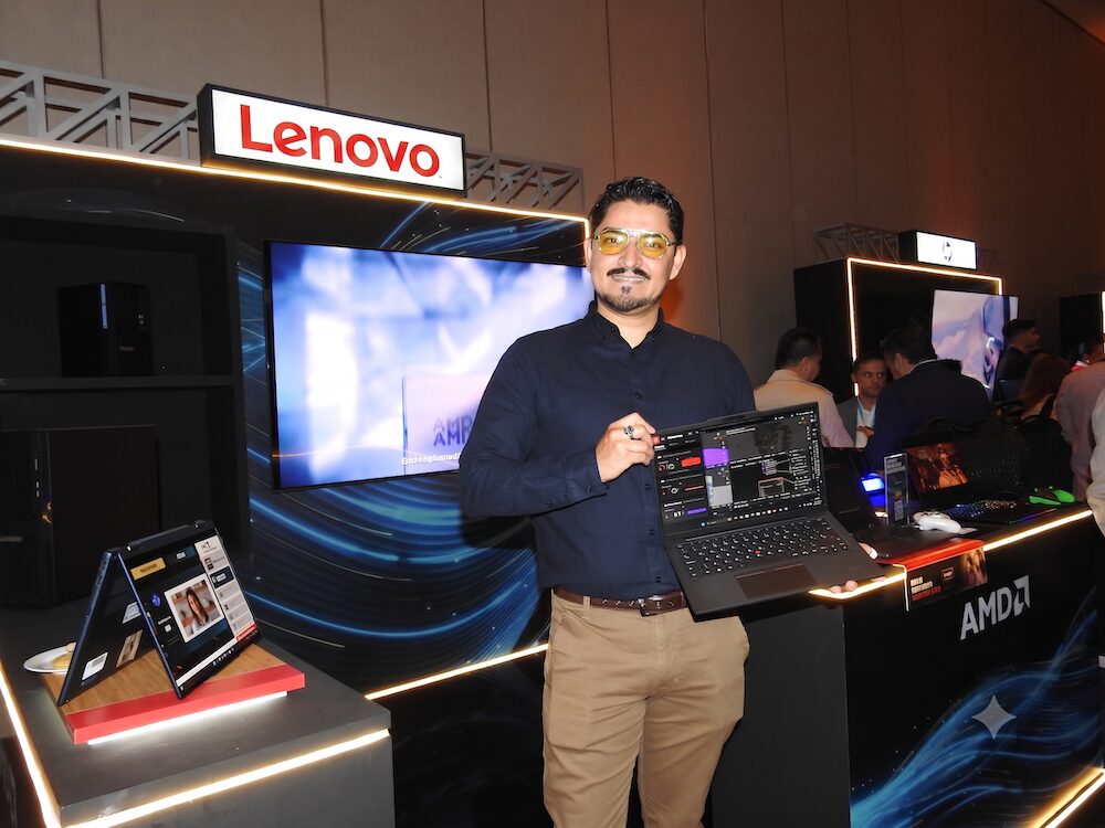 Hombre con lentes amarillos y camisa oscura sosteniendo una laptop Lenovo ThinkPad abierta con una sola mano. Se encuentra en el stand de exhibición de Lenovo, rodeado de pantallas, dispositivos y luces decorativas del evento.