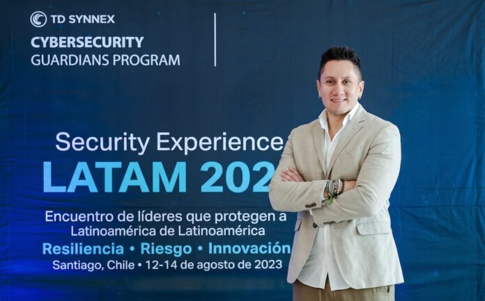 Fotografía de Uriel Sánchez posando de pie con los brazos cruzados; viste una camisa blanca y un saco beige, con un fondo azul de TD SYNNEX y Cisco que enmarca el evento Security Experience LATAM.