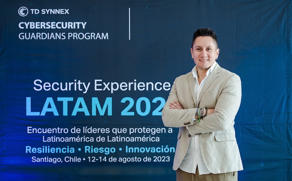 Fotografía de Uriel Sánchez posando de pie con los brazos cruzados; viste una camisa blanca y un saco beige, con un fondo azul de TD SYNNEX y Cisco que enmarca el evento Security Experience LATAM.
