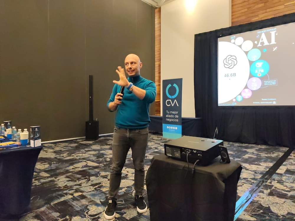 Fotografía de Engel Fonseca hablando con un micrófono en la mano y gesticulando, de pie junto a un proyector. De fondo, una pantalla muestra una diapositiva con el título "AI" y logotipos relacionados con inteligencia artificial.