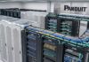 Panduit fortalece la infraestructura de conectividad para el regreso a clases Fotografía de un cuarto de telecomunicaciones moderno e iluminado que muestra múltiples racks y gabinetes de la marca Panduit. Destacan gruesos mazos de cables de red en colores azul, amarillo y verde, los cuales están peinados y organizados de manera impecable sobre bandejas horizontales. A la izquierda se aprecian pasillos contenidos con techos transparentes, y al fondo resalta un gran logotipo negro de Panduit montado sobre un panel de acabado metálico.