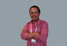 DACAS acelera su estrategia alrededor de los datos, el canal y la IA Fotografía de retrato de Fernando López, country manager de DACAS para Colombia y México, posando de frente con los brazos cruzados y sonriendo a la cámara. Viste una camisa rosa de manga larga remangada y lleva un gafete de acreditación al cuello, sobre un fondo gris claro uniforme.