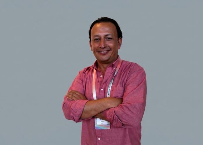 Fotografía de retrato de Fernando López, country manager de DACAS para Colombia y México, posando de frente con los brazos cruzados y sonriendo a la cámara. Viste una camisa rosa de manga larga remangada y lleva un gafete de acreditación al cuello, sobre un fondo gris claro uniforme.