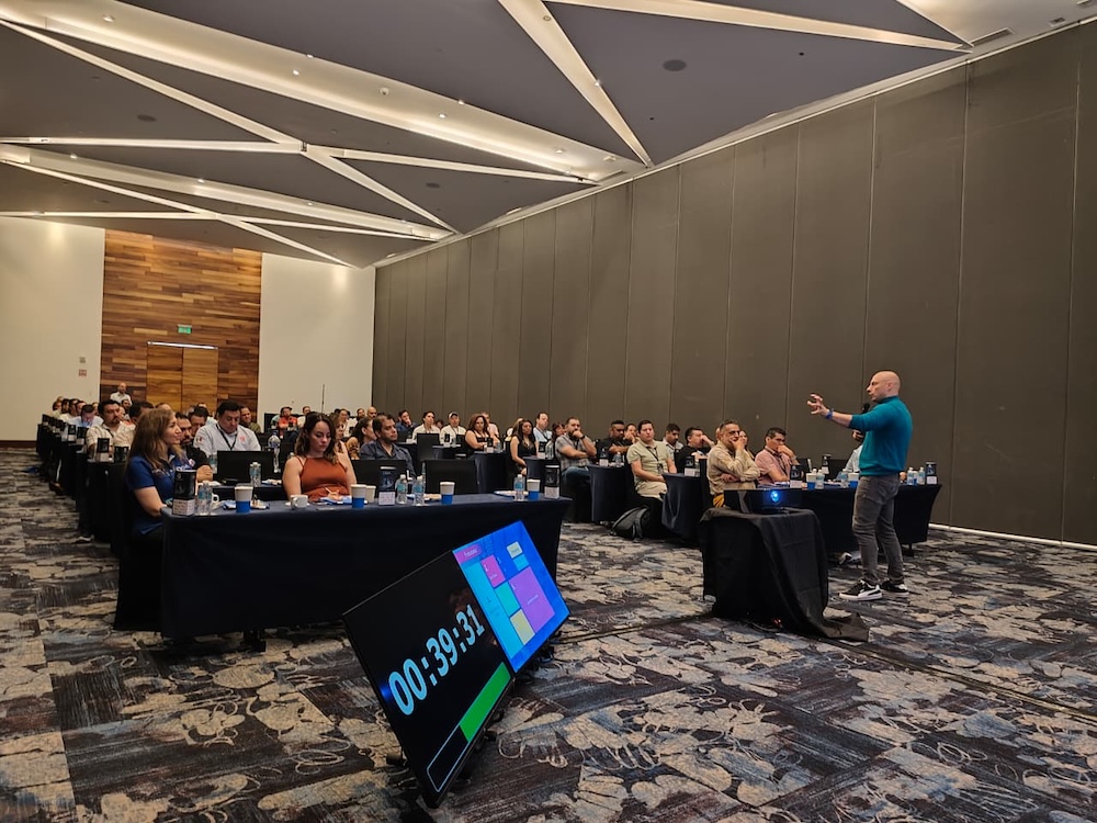 Fotografía de un hombre calvo con suéter azul impartiendo una conferencia ante una audiencia atenta en un amplio salón. En primer plano destaca un monitor en el suelo mostrando un temporizador en cuenta regresiva.