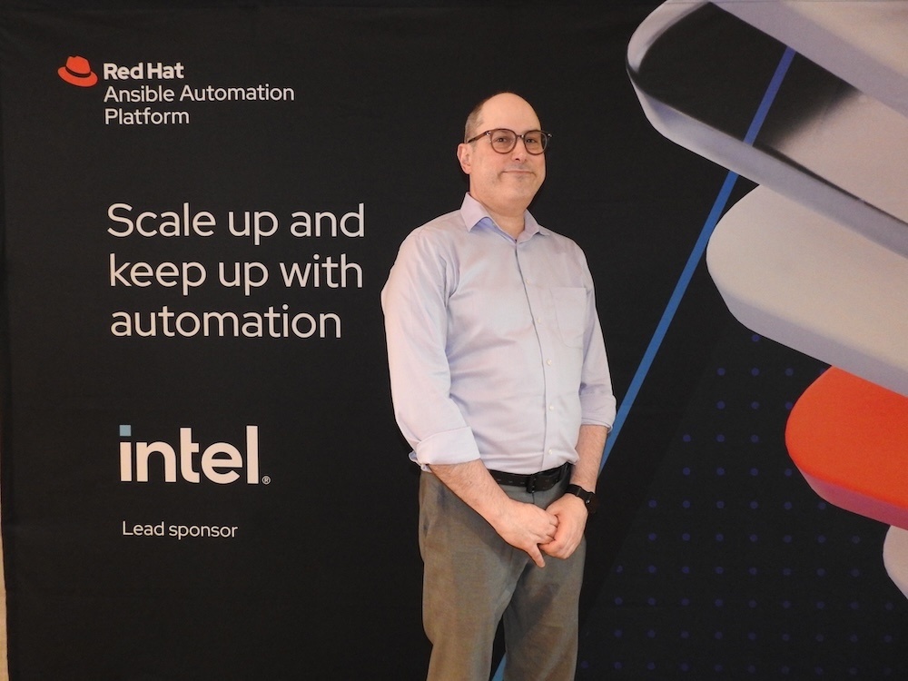 Automatización de TI con Red Hat Ansible: Fotografía de Chad Ferman posando de frente con las manos entrelazadas a la altura de la cintura, vistiendo una camisa azul claro de botones y pantalones grises. Se encuentra frente a un muro promocional oscuro de Red Hat Ansible Automation Platform.