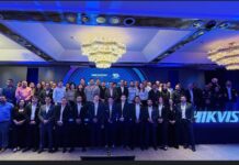Hikvision impulsa la seguridad inteligente con IA, AIoT y ciberseguridad Fotografía grupal de numerosos asistentes posando de pie sobre un escenario; al fondo se observa una gran pantalla con el logotipo de Hikvision y gráficos del evento bajo una iluminación en tonos azules y grandes candelabros en el techo.