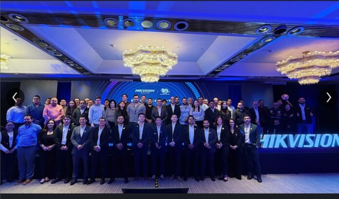 Fotografía grupal de numerosos asistentes posando de pie sobre un escenario; al fondo se observa una gran pantalla con el logotipo de Hikvision y gráficos del evento bajo una iluminación en tonos azules y grandes candelabros en el techo.
