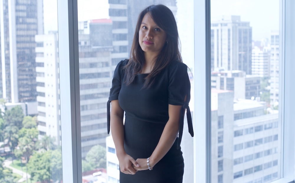 Fotografía de Arely Reyes, analista de Select, posando de pie con un vestido negro de manga corta frente a un gran ventanal que muestra edificios de la ciudad.
