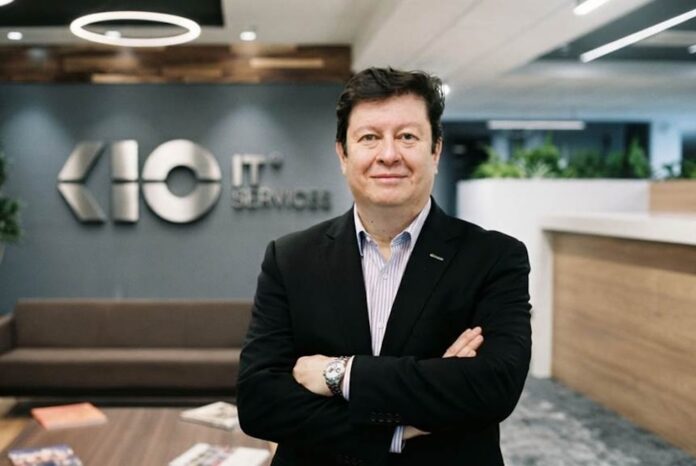 Fotografía de Rafa Sánchez Loza, quien asume el cargo de Chief Revenue Officer, posando de pie con los brazos cruzados vistiendo un saco oscuro y camisa clara. Se encuentra en unas instalaciones corporativas modernas, con el logotipo metálico de KIO IT Services visible en la pared del fondo.