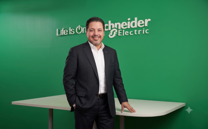 La evolución del canal TI. De proveedores de hardware a consultores estratégicos Fotografía de José Alberto Llavot, Gerente de Preventa y Desarrollador de Negocios en Schneider Electric, posando de pie y sonriendo a la cámara. Viste un traje oscuro con camisa blanca sin corbata, apoyando una mano sobre una mesa blanca frente a una pared verde brillante que exhibe el logotipo tridimensional de la compañía con el lema