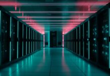 Janus Henderson: Centros de datos sedientos; cómo la IA está redefiniendo la demanda y el riesgo del agua Pasillo simétrico de un centro de datos moderno con filas de servidores oscuros a ambos lados, resaltados por franjas de luz LED cian en los gabinetes y tubos de iluminación de neón rosa en el techo.