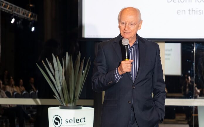 La inteligencia artificial como catalizador del modelo de negocio Fotografía de Ricardo Zermeño, director general de Select, hablando por un micrófono durante un evento, vistiendo un saco oscuro y camisa a rayas, con una planta y el logotipo de su empresa visibles en primer plano.