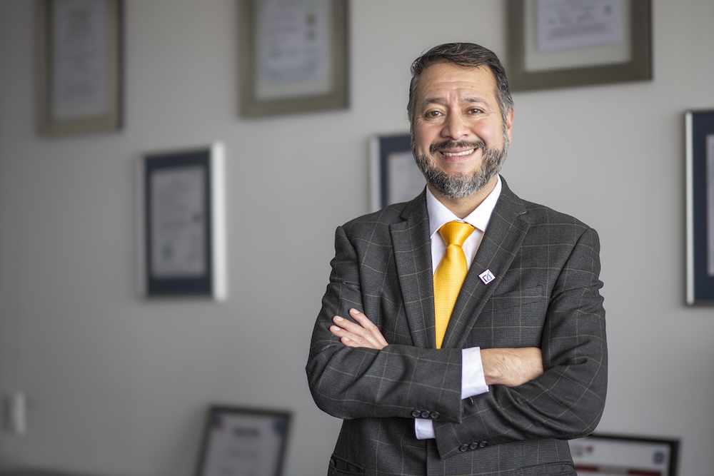Retrato de Julio Olmos, un hombre con barba entrecana, vistiendo un saco gris oscuro a cuadros, camisa blanca y una corbata amarilla vibrante. Posa con los brazos cruzados y una sonrisa frente a una pared decorada con diversos certificados enmarcados.