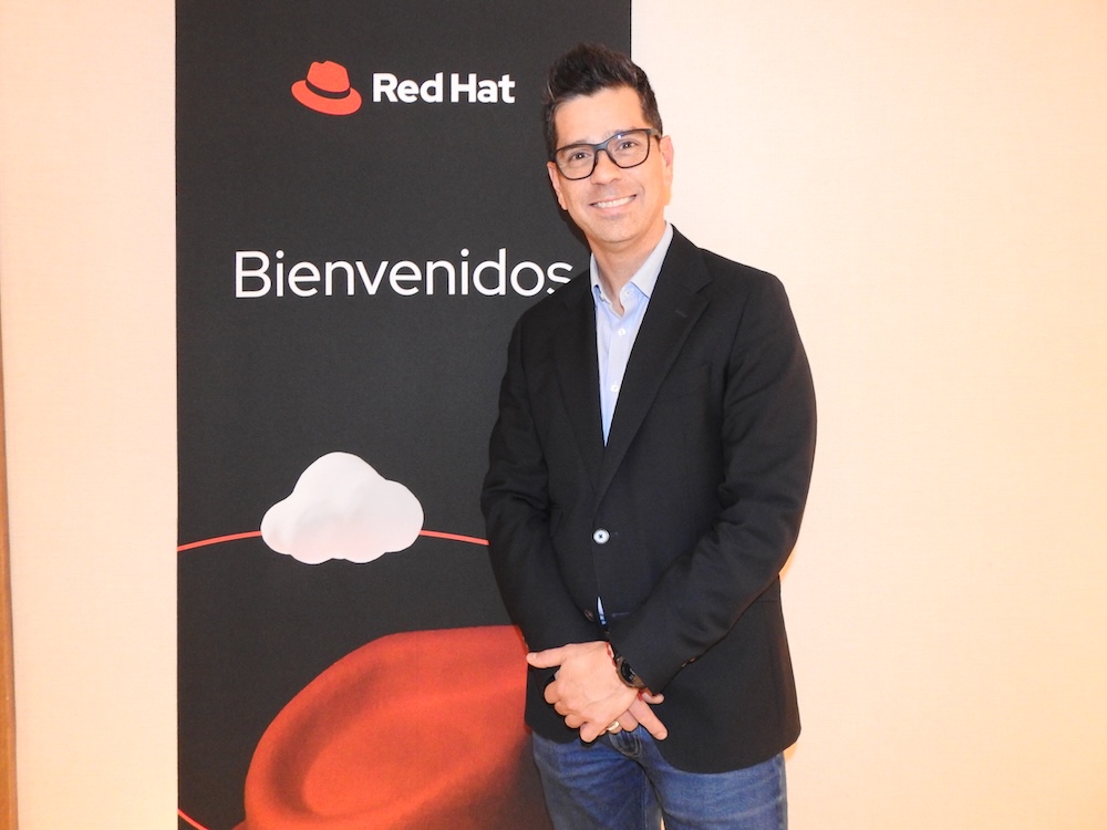 Retrato fotográfico de Daniel Romero sonriendo a la cámara, con las manos entrelazadas al frente. Viste un saco oscuro sobre una camisa clara y jeans. Detrás de él hay un panel negro con el logotipo de Red Hat y la palabra "Bienvenidos".
