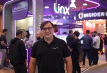 Totvs Napse: Digitalización del retail impulsa la eficiencia y el crecimiento de las PyMEs Fotografía de Martín Malievac sonriendo a la cámara, usando anteojos y vistiendo una camisa negra tipo polo. Se encuentra de pie en los pasillos de una feria corporativa o evento tecnológico, con un stand de exhibición iluminado con luces de neón moradas desenfocado en el fondo.
