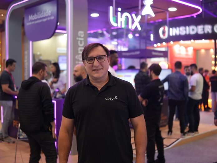 Fotografía de Martín Malievac sonriendo a la cámara, usando anteojos y vistiendo una camisa negra tipo polo. Se encuentra de pie en los pasillos de una feria corporativa o evento tecnológico, con un stand de exhibición iluminado con luces de neón moradas desenfocado en el fondo.