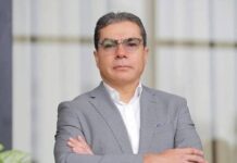 Worldpanel by Numerator revela que el pago con tarjeta eleva 22% el ticket en tienditas Fotografía de un hombre adulto con anteojos y cabello corto peinado hacia atrás, que mira fijamente a la cámara. Viste un traje gris claro sobre una camisa blanca sin corbata y mantiene los brazos cruzados. El fondo es un entorno arquitectónico difuminado con iluminación natural.