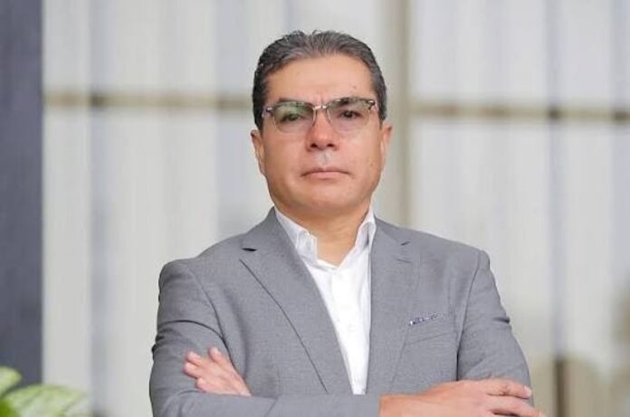 La transformación del canal tradicional ante un consumidor más exigente Fotografía de un hombre adulto con anteojos y cabello corto peinado hacia atrás, que mira fijamente a la cámara. Viste un traje gris claro sobre una camisa blanca sin corbata y mantiene los brazos cruzados. El fondo es un entorno arquitectónico difuminado con iluminación natural.