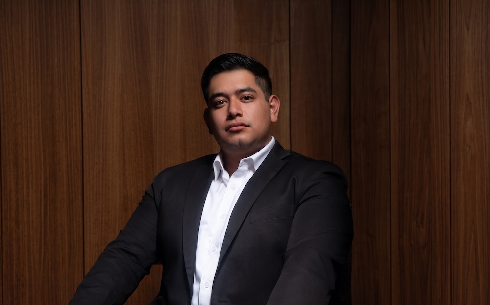 Retrato de Alan Adolfo López, analista de Select, vistiendo un saco negro y camisa blanca sin corbata, sentado frente a un fondo de paneles de madera oscura.