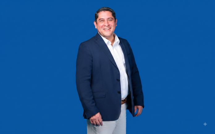 Fotografía de medio cuerpo de Víctor Urzúa, quien ocupa el cargo de subdirector comercial en CompuSoluciones. En la imagen, el ejecutivo aparece sonriendo hacia la cámara, vistiendo una camisa blanca sin corbata y un saco oscuro, posando frente a un fondo liso de color azul.