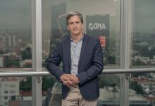 AMD potencia la IA y redefine la infraestructura tecnológica con una oportunidad de 2 billones de dólares Fotografía de Nicolás Cánovas, un hombre de cabello entrecano vistiendo un saco azul oscuro sobre una camisa clara. Posando con las manos entrelazadas frente a un amplio ventanal que refleja el logotipo de AMD y muestra un paisaje urbano nublado.
