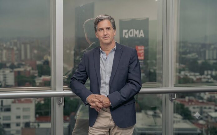 La visión estratégica de AMD para la inteligencia artificial en América Latina Fotografía de Nicolás Cánovas, un hombre de cabello entrecano vistiendo un saco azul oscuro sobre una camisa clara. Posando con las manos entrelazadas frente a un amplio ventanal que refleja el logotipo de AMD y muestra un paisaje urbano nublado.