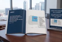 Xerox es reconocida como líder mundial en soluciones para salas de correo Fotografía de una mesa de conferencias de madera sobre la cual descansan un reporte impreso abierto y un letrero de acrílico transparente. Ambos elementos exhiben el reconocimiento de Xerox en el cuadrante de "Líderes" dentro del gráfico de IDC MarketScape de 2026. De fondo, se observan amplios ventanales con vista a edificios de la ciudad.