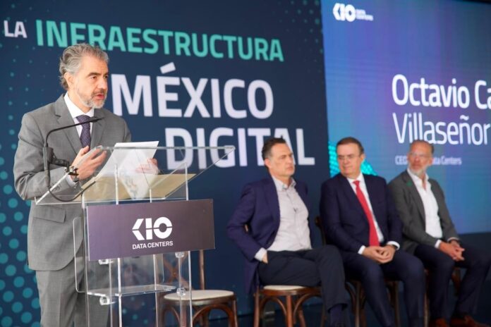 Fotografía de Octavio Camarena, director general de KIO Data Centers, hablando desde un atril de acrílico transparente. Al fondo, el secretario de Economía Marcelo Ebrard y otros dos hombres lo escuchan sentados frente a una gran pantalla con el nombre del orador y un panel con el lema 
