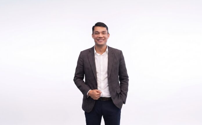Fotografía de medio cuerpo de Luis Nogales, sonriendo a la cámara, vistiendo un saco oscuro texturizado y una camisa blanca sin corbata, sobre un fondo completamente blanco.