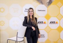 Axis Communications: La inteligencia artificial llega a las ciudades, tecnología para anticipar riesgos y mejorar la seguridad Fotografía de retrato de Diana Ardila, sonriendo a la cámara mientras apoya un brazo sobre el respaldo de una silla blanca. Viste un traje sastre negro con una blusa oscura de puntos traslúcida y posa frente a un fondo corporativo amarillo y blanco decorado con hexágonos, íconos tecnológicos y el logotipo de Axis Communications.