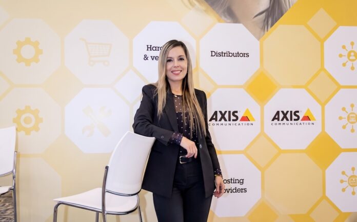 Liderazgo estratégico en el desarrollo tecnológico de ciudades conectadas Fotografía de retrato de Diana Ardila, sonriendo a la cámara mientras apoya un brazo sobre el respaldo de una silla blanca. Viste un traje sastre negro con una blusa oscura de puntos traslúcida y posa frente a un fondo corporativo amarillo y blanco decorado con hexágonos, íconos tecnológicos y el logotipo de Axis Communications.