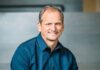 Dynatrace adquiere Arize por 915 millones de dólares para reforzar su observabilidad de IA Fotografía de medio cuerpo de Rick McConnell, sonriendo a la cámara y vistiendo una camisa azul oscura, con un fondo arquitectónico desenfocado en tonos grises.