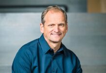 Dynatrace adquiere Arize por 915 millones de dólares para reforzar su observabilidad de IA Fotografía de medio cuerpo de Rick McConnell, sonriendo a la cámara y vistiendo una camisa azul oscura, con un fondo arquitectónico desenfocado en tonos grises.