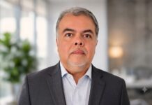 Red Hat fortalece su estrategia tecnológica en América Latina con nuevo Chief Architect, Sinuhé Sánchez Fotografía de retrato de Sinuhé Sánchez. Es un hombre de mediana edad, de tez clara, con cabello entrecano y barba de candado. Viste un saco de traje color gris oscuro sobre una camisa de vestir clara con un sutil patrón de cuadros. Se encuentra de frente a la cámara con una expresión neutra y profesional. El fondo está completamente desenfocado, apreciándose un entorno de oficina moderna bien iluminada con una planta verde a la izquierda.