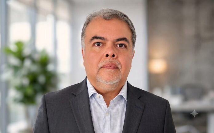 Liderazgo tecnológico. Sinuhé Sánchez asume como Chief Architect de Red Hat para el norte de Latinoamérica Fotografía de retrato de Sinuhé Sánchez. Es un hombre de mediana edad, de tez clara, con cabello entrecano y barba de candado. Viste un saco de traje color gris oscuro sobre una camisa de vestir clara con un sutil patrón de cuadros. Se encuentra de frente a la cámara con una expresión neutra y profesional. El fondo está completamente desenfocado, apreciándose un entorno de oficina moderna bien iluminada con una planta verde a la izquierda.