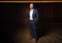 Logicalis fortalece su crecimiento en NOLA con el nombramiento de Adrián Moheno Fotografía de cuerpo entero de Adrián Moheno sonriendo a la cámara. Lleva anteojos transparentes, un traje azul marino, camisa blanca sin corbata, cinturón café y zapatos de cuero en tono marrón. Posando de pie con las manos en los bolsillos del pantalón, se encuentra sobre un piso de madera en un espacio de fondo oscuro con iluminación cálida superior.