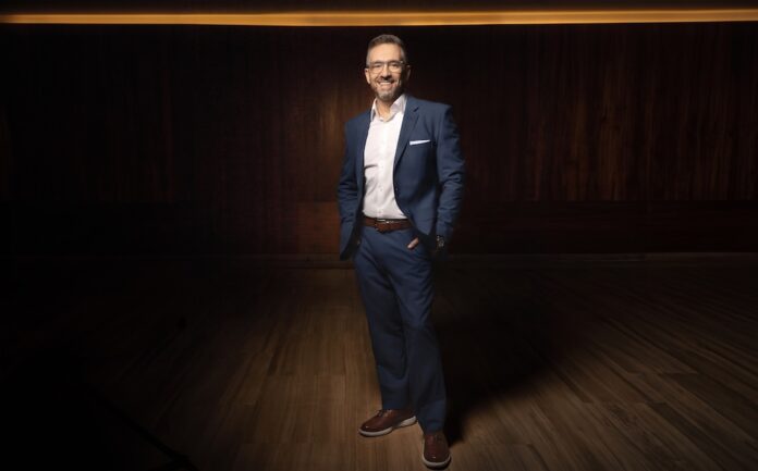 Logicalis refuerza su liderazgo regional con el nombramiento de Adrián Moheno Fotografía de cuerpo entero de Adrián Moheno sonriendo a la cámara. Lleva anteojos transparentes, un traje azul marino, camisa blanca sin corbata, cinturón café y zapatos de cuero en tono marrón. Posando de pie con las manos en los bolsillos del pantalón, se encuentra sobre un piso de madera en un espacio de fondo oscuro con iluminación cálida superior.