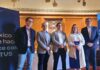 TOTVS fortalece a México como hub estratégico para su expansión en América Latina Fotografía grupal de cinco ejecutivos de TOTVS, cuatro hombres y una mujer, posando de pie y sonriendo a la cámara en un salón clásico con paredes revestidas en madera y piso de duela. A la izquierda se observa parcialmente un banner azul con el logotipo corporativo de la empresa.