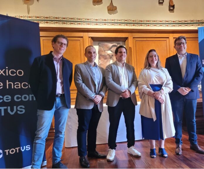 Fotografía grupal de cinco ejecutivos de TOTVS, cuatro hombres y una mujer, posando de pie y sonriendo a la cámara en un salón clásico con paredes revestidas en madera y piso de duela. A la izquierda se observa parcialmente un banner azul con el logotipo corporativo de la empresa.